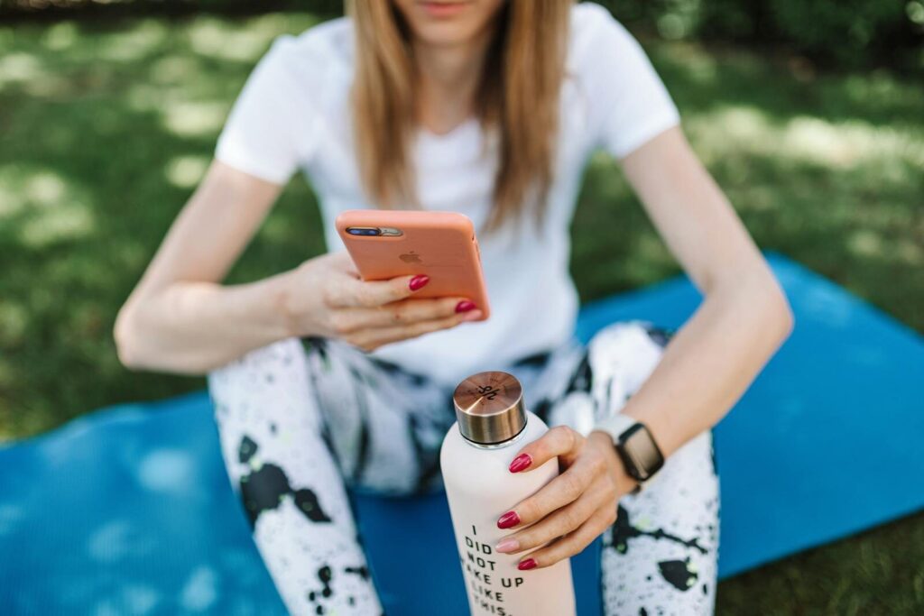 Woman using smartphone while holding bottle on yoga mat outdoors.